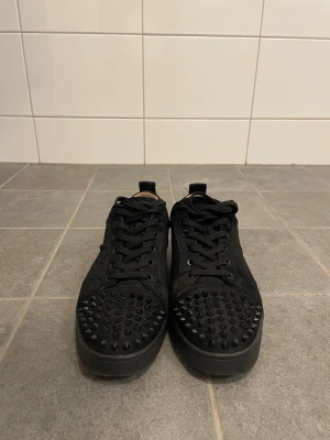 Svarta Louboutin  - Svarta sneakers med rund tå och platt sula, täckta med svarta nitar på tån för en edgy look. Skorna har snörning och är gjorda i mocka med beige innersida. Perfekta för dig som gillar att sticka ut med detaljer.