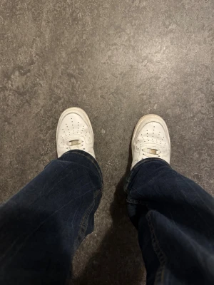 Nike Air Force 1 - Klassiska Nike Air Force 1 sneakers i helvitt skinn med perforerad tå och tydlig AF1-detalj på snörningen. Snygg broderad Nike Air-logga på hälen och platt sula. Perfekta för dig som gillar en clean och tidlös look.