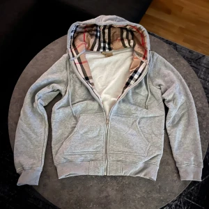 Grå Burberry Hoodie - Snygg grå hoodie från Burberry i storlek M med klassiskt rutigt foder i huvan. Tröjan har dragkedja framtill, två fickor och ribbade muddar. Tillverkad i mjuk bomull med lite elastan för extra komfort. Perfekt för dig som gillar stilrena och exklusiva plagg. 