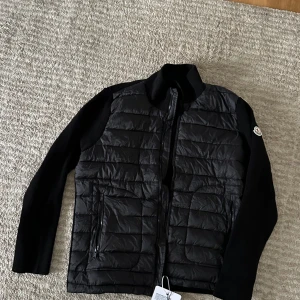 Svart quiltad pufferjacka Moncler - Svart pufferjacka från Moncler med quiltad front och stickade ärmar. Jackan har dragkedja framtill, två sidofickor med dragkedja och Moncler-logga på ärmen. Snygg, stilren design med hög krage och en mix av material som ger en cool look.nfc tag