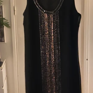 Svart långklänning med paljetter Fransa - Svart långklänning från Fransa med snyggt paljettband i silver och guldtoner längs hela framsidan. Ärmlös modell med rund halsringning och rak passform. Perfekt för dig som vill sticka ut med lite extra bling.