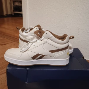 Reebok Royal Prime Mid 2.0 sneakers - Snygga Reebok Royal Prime Mid 2.0 sneakers i vitt med bruna detaljer och gul logga. Skorna har klassisk snörning, mellanhögt skaft och är tillverkade i syntetmaterial med mjuk vadderad insida. Perfekta för dig som gillar stilrena och sportiga vibes. Barnskor