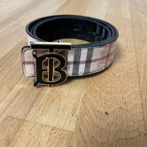 Burberry rutig bälte med TB-spänne - Säljer ett Burberry bälte med ikoniskt TB-spänne i metall. Ena sidan har klassiskt beige, svart och rött rutmönster, andra sidan är svart med diskret rutig struktur. Bältet är tillverkat i skinn och har en lyxig känsla med tydliga Burberry-vibbar.