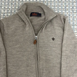 Beige kofta i merinoull från Morris - Snygg beige kofta från Morris i 100% merinoull. Tröjan har hel dragkedja med läderdetalj, ribbade muddar och ett diskret Morris-emblem broderat på bröstet. Perfekt för dig som gillar stilrena och klassiska plagg med premiumkänsla. Storlek m men sitter mer som s/xs