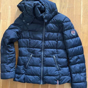 Tommy Hilfiger jacka - Snygg mörkblå dunjacka från Hilfiger Denim med huva och dragkedja framtill. Jackan har två snedställda fickor med dragkedja och ett märke på ena ärmen. Quiltad design som håller värmen under kalla dagar.
