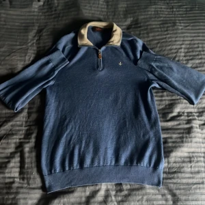 Blå merinoulltröja Morris half zip - Snygg blå tröja från Morris i 100% merinoull med beige krage och half zip-dragkedja. Tröjan har ribbade muddar, kort ärm och en liten broderad logga på bröstet. Perfekt för dig som gillar klassisk stil med en modern touch.