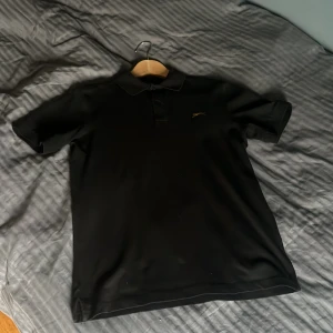 Svart pikétröja från Slazenger S - Svart pikétröja från Slazenger i storlek S. Klassisk design med krage, korta ärmar och två knappar framtill. Tröjan har en broderad guldfärgad panter-logga på bröstet. Materialet är mjuk och stretchig bomull med 5% elastan för extra komfort.