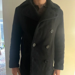 Svart skepparkavaj med bred krage - Säljer en svart skepparkavaj med bred krage och dubbelknäppning framtill. Jackan har två sidofickor och är tillverkad i ett tjockt ullmaterial som håller dig varm. Passformen är normal och den har långa ärmar. Perfekt för dig som gillar klassisk stil med en modern touch.