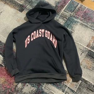 Svart US Coast Guard hoodie - Svart hoodie med stor vit och röd US Coast Guard-text på bröstet. Klassisk huva och magficka framtill, ribbade muddar vid ärmslut och nederkant. Perfekt för dig som gillar college-vibe och vill ha en enkel, stilren look.