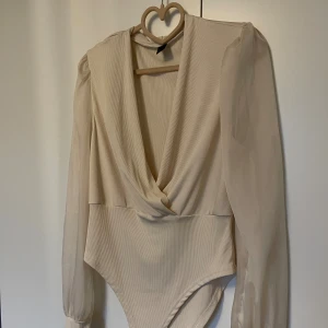 Beige omlott bodysuit med puffärm - Säljer en beige bodysuit med omlott-drapering framtill och djup v-ringning. Toppen har långa puffärmar i transparent material och ribbad struktur på kroppen. Perfekt för dig som gillar stilrena och trendiga plagg med extra detaljer.helt ny aldrig använd