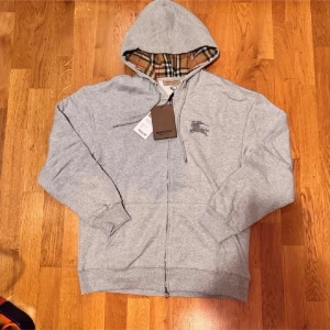 Grå Burberry hoodie med dragkedja - Grå hoodie från Burberry med klassiskt rutigt foder i huvan och broderad logga på bröstet. Tröjan har dragkedja framtill, snörning i huvan och ribbade muddar. Perfekt för dig som vill ha en stilren och lyxig streetwear-look.