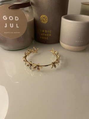 Guldfärgat armband med blommor - Superfint armband i guldfärg med detaljerade blommor runt hela bandet. Armbandet har en öppen design och blommorna ger en romantisk och trendig känsla. Perfekt accessoar för att lyfta din outfit.