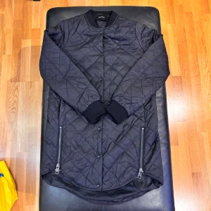 Svart quiltad jacka från Vero Moda - Quiltad svart jacka från Vero Moda i storlek S. Jackan har lång ärm, ribbad krage och muddar, samt två dragkedjor framtill. Den har en längre, rundad nederkant och knäpps med knappar. Perfekt för dig som gillar stilren och enkel design.