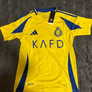 Al Nassr gul/blå fotbollströja Adidas - Säljer en officiell Al Nassr fotbollströja från Adidas i slim fit. Tröjan är gul med blå detaljer, klubbmärke och sponsortryck på bröstet. Materialet är lätt och ventilerande polyester, perfekt för match eller träning. Klassisk V-ringning och korta ärmar.
