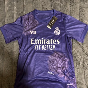 Real Madrid Y-3 lila fotbollströja - Exklusiv Real Madrid 120-årsjubileumströja från Y-3 i lila polyester med blommigt mönster. Kortärmad modell med rund hals, tryckt klubbmärke och Emirates Fly Better-logga på bröstet. Snygg design med rosor och Y-3 branding.