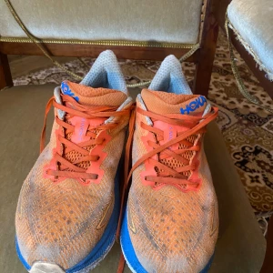 Hoka Clifton 9 orange/blå sneakers - Säljer ett par Hoka Clifton 9 sneakers i orange med blå detaljer och vita inslag. Skorna har mesh-ovandel för bra ventilation, platt sula och snörning. Perfekta för löpning eller träning, med sportig look och bekväm passform. Snyggt Hoka-tryck på hälen och sidan.