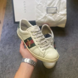 Priset kan sänkas!!!(Gucci Ace (allt OG)  - Gucci Ace sneakers i vitt skinn med klassiska gröna och röda ränder på sidan och broderad guldfärgad bi. Baksidan har metallicröd och metallicgrön hälflik med textur. Platt sula, rund tå och snörning. Ikonisk och stilren modell med lyxig känsla.