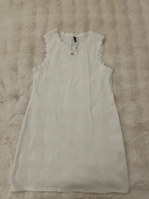 Vit ärmlös klänning från H&M - Supersöt vit ärmlös klänning från H&M med vågiga kanter vid ärmhålen. Enkel och stilren design i mjuk bomull, perfekt för varma dagar. Rak passform och kort längd gör den både bekväm och trendig. Klänningen går att tvätta för att få bort sminkfläckarna. Aldrig använd utan köptes så!