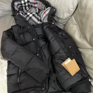 Svart pufferjacka från Burberry - Svart pufferjacka från Burberry med klassisk rutig huva i grått, vitt och rött. Jackan har dragkedja och knappar framtill, två sidofickor med dragkedja och en quiltad design. Perfekt för kalla dagar med sin tjocka och värmande stil. [storlek L men skulle säga passar mer till S/M