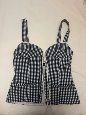 Svartvit rutig korsettopp från Zara - Säljer en snygg korsettopp från Zara i svartvitt rutigt mönster. Toppen har breda axelband, snörning framtill och markerade kupor för en cool siluett. 