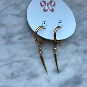 Guldiga örhängen  - Snygga örhängen från WOS i guldtonad metall. De har rundade ringar med kedjor som avslutas med en spetsig, avlång hängande detalj. Perfekt för dig som gillar edgy och trendiga accessoarer med lite attityd.
