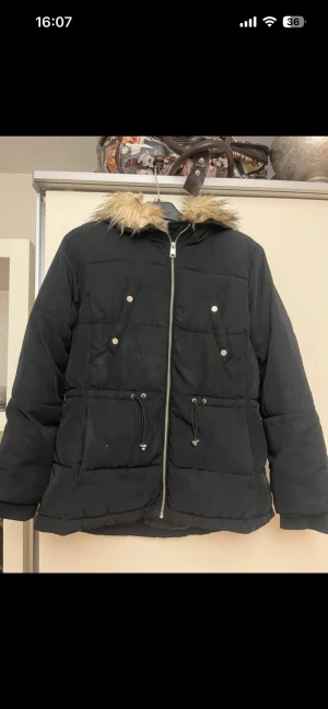 Svart pufferjacka med fuskpäls - Svart pufferjacka med fluffig fuskpäls på huvan och snygga snörningsdetaljer i midjan. Jackan har dragkedja framtill, två stora fickor med knappar och en skön, lite loose passform. Perfekt för kalla dagar när du vill vara både varm och stilren.