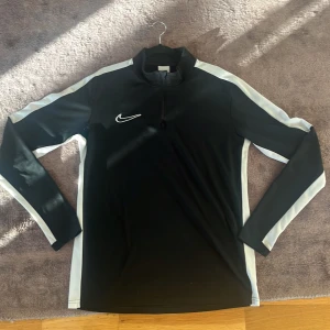 Nike svart/vit träningströja med halvzip - En stilren och sportig långärmad träningströja från Nike. Den är svart med vita paneler längs armarna och sidorna, samt har en halv dragkedja framtill. Materialet ser ut att vara stretchigt och anpassat för fysisk aktivitet. Perfekt för fotbollsträning, löpning eller vardagligt sportigt bruk.  i fint skick.