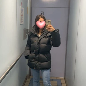 Bommerang jacka fin skick xs - Säljer en svart pufferjacka med stor huva och praktiska fickor. Jackan har knappar och dragkedja framtill, samt en quiltad design som ger extra värme. Perfekt för kalla dagar och har en snygg, lite oversized passform.