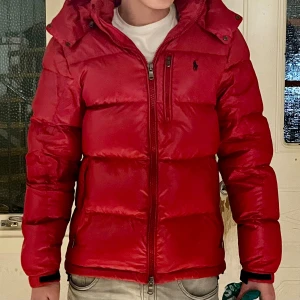 Röd pufferjacka från Polo Ralph Lauren - Säljer en röd pufferjacka från Polo Ralph Lauren med huva och svart logga på bröstet. Jackan har dragkedja framtill, sidofickor och en extra bröstficka med zip. Materialet är glansigt och jackan är riktigt dunig och varm, perfekt för kalla dagar. Modellen är Quilted recycled shell storlek small, nypris ca 6000 och uppåt. Sparsamt använd. Hund finns i hemmet.