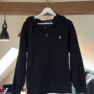 Svart hoodie från Polo Ralph Lauren | Hoodie - Svart hoodie från Polo Ralph Lauren med dragkedja och huva. Klassisk design med vit broderad logga på bröstet. Tvär snygg och funkar till allt verkligen. Storlek M passar nog bra på någon med S också. Mvh Anton