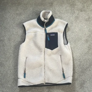 Vit fleeceväst från Patagonia - Säljer en vit fleeceväst från Patagonia med marinblå detaljer och dragkedjor. Västen har hög krage, tre fickor med dragkedja och ett mjukt, fluffigt material som håller dig varm. Perfekt för lager-på-lager och outdoor-häng.