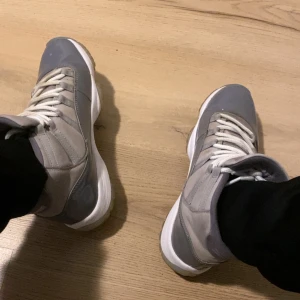 Nike Air Jordan 11 Cool Grey High - Nike Air Jordan 11 Cool Grey highs med grå och ljusgrå färgblock, vit sula och vita snören. Skorna har en blank patentläderpanel, mesh och mockadetaljer samt Jumpman-logga på hälen. Klassisk rund tå och platt sula för en clean look.