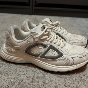 Vita D sneakers  - Säljer ett par snygga sneakers i storlek 41 i vit med gråa detaljer och tydlig CD-logga på sidan. Skorna har rund tå, platt sula och snörning. Ovandelen är i en mix av skinn och mesh för en cool och sportig vibe. Perfekta för dig som gillar exklusiva märken och streetstyle.