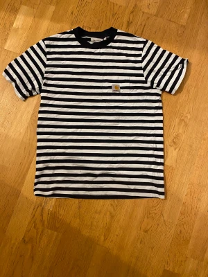 Randig Carhartt t-shirt svart/vit - Snygg randig t-shirt från Carhartt i svart och vitt med klassisk logga på bröstfickan. Modellen har rund hals och korta ärmar, tillverkad i mjuk bomull. Perfekt för dig som gillar streetwear!! Den är i storlek S men passar som M. Den är nästan helt ny. Jag har använt den 2 gånger!