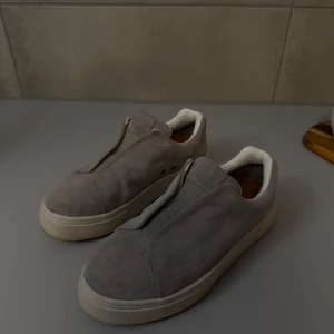 Eytys Doja Grå Mocka - Säljer ett par grå slip-on sneakers från Eytys i mocka med vit innersida och platt sula. Skorna har en stilren design utan snörning, elastisk panel framtill och en lila detalj på hälen. Perfekt för dig som gillar minimalistisk och clean stil.