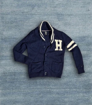 Marinblå kofta från Hilfiger Denim - Snygg marinblå kofta från Hilfiger Denim med vita ränder på ena ärmen och stor H på bröstet. Hör av dig vid frågor!