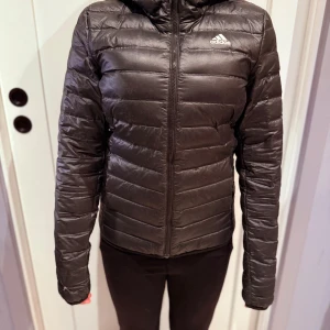 Svart pufferjacka från Adidas XS - Svart pufferjacka från Adidas med huva och dragkedja framtill. Jackan har horisontella quiltade paneler och Adidas-logga på bröstet. Långärmad och med en slim passform, perfekt för kyliga dagar. Materialet är syntet och jackan är lätt och smidig. Finns en väldigt liten lagning på armmudden, se bild. 