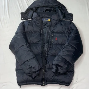 Svart pufferjacka från Polo Ralph Lauren - Svart pufferjacka från Polo Ralph Lauren med avtagbar huva, dragkedja framtill och röd broderad logga på bröstet. Jackan har quiltad design och flera praktiska fickor med dragkedja. Perfekt för kalla dagar och har en riktigt clean look.