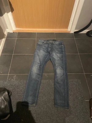 Tiger of Sweden Pistolero jeans blå 29/30 - Snygga blå jeans från Tiger of Sweden i modellen Pistolero. Klassisk femficksdesign med orangea sömmar och svart läderpatch bak. Jeansen har rak passform och är tillverkade i slitstarkt denim. Perfekt för dig som gillar stilrena och tidlösa jeans.