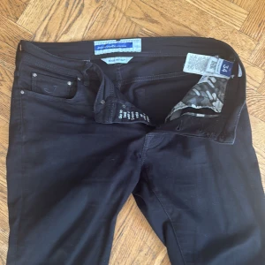 Svarta jeans från Jacob Cohen, stl 34 - Snygga svarta jeans från Jacob Cohen med klassisk femficksdesign och diskret logga på myntfickan. Byxorna har smal passform och är tillverkade i ett stretchigt bomullsmaterial. Insidan av linningen har ett camomönstrat tyg för en cool detalj.