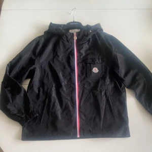Svart vindjacka från Moncler - Stilren svart vindjacka från Moncler med en iögonfallande dragkedja i rött, vitt och blått. Lätt och funktionell jacka – perfekt för blåsiga dagar och övergångssäsong. | Storlek: M / 2, Färg: Svart, Detaljer: Trikolor-dragkedja, bröstficka med Moncler-logga, Fickor: Två sidofickor med dragkedja, Skick: Bra | Tidlös design med tydliga Moncler-detaljer. Enkel att styla och funkar både till vardag och mer uppklädda outfits. | Pris: Kan diskuteras | Skickas snabbt | 