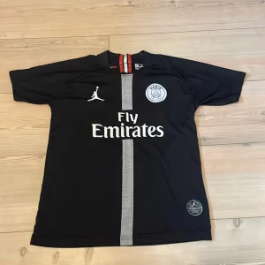 PSG Neymar Jr svart fotbollströja - Svart PSG fotbollströja med Neymar Jr och nummer 10 på ryggen. Framsidan har Fly Emirates-tryck, PSG-emblem och Jordan-logga. Tröjan har en vertikal rand i grått och röda detaljer vid halsen. Tillverkad i lätt, ventilerande material och har korta ärmar.