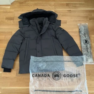 Svart dunjacka från Canada Goose - Säljer en mörk grå dunjacka från Canada Goose med avtagbar huva och pälsdetalj. Jackan har en stilren look med diskret logga på ärmen, dragkedja och knappar framtill samt praktiska fickor. Perfekt för kalla dagar och riktigt snygg till streetwear.