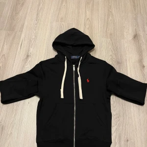 Svart hoodie från Polo Ralph Lauren - Svart hoodie från Polo Ralph Lauren med vit snörning och dragkedja framtill. Klassisk röd broderad logga på bröstet och huva. Tröjan har långa ärmar och är gjord i mjuk bomull, perfekt för chill dagar. Fickor framtill för extra casual vibe.