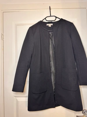 Svart rock från H&M, storlek 38 - Stilren svart rock från H&M i storlek 38. Jackan har rundad hals, två stora fickor framtill och är helfodrad. Materialet är strukturerat och känns mjukt mot huden. Perfekt för dig som gillar minimalistisk och clean look.