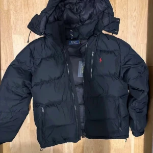 Svart pufferjacka från Polo Ralph Lauren - Svart pufferjacka från Polo Ralph Lauren med avtagbar huva, röda logobroderiet på bröstet och flera dragkedjeförsedda fickor. Jackan har en quiltad design och är tillverkad i syntetmaterial som håller dig varm under kalla dagar. Perfekt för dig som gillar stilren och sportig look.