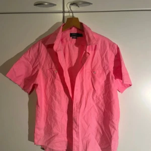 Rosa kortärmad skjorta Polo Ralph Lauren - Fräsch rosa skjorta från Polo Ralph Lauren i klassisk passform. Skjortan har korta ärmar, krage och knappar framtill samt två bröstfickor med knapp. Tillverkad i mjuk bomull, perfekt för varma dagar. Snygg och enkel att matcha med jeans eller shorts.