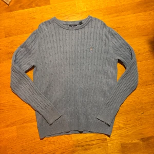 Ljusblå kabelstickad tröja från GANT - Snygg ljusblå kabelstickad tröja från GANT med broderad logga på bröstet. Tröjan har rund hals, ribbade muddar och långärmad passform. Perfekt för dig som gillar klassisk stil med en modern touch.