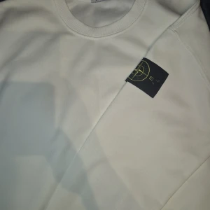 Vit sweatshirt från Stone Island - Vit långärmad sweatshirt från Stone Island med klassisk rund hals och den ikoniska svarta patchen med gul broderad logga på vänster ärm. Tröjan har ribbade muddar och är tillverkad i mjuk bomull för skön komfort. Perfekt för dig som gillar clean och stilren streetwear.
