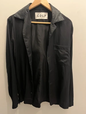 Svart satin skjorta från CDLP - Svart skjorta från CDLP i mjuk satin med klassisk krage och bröstficka. Skjortan har en clean look med knappar framtill och lång ärm. Perfekt för dig som gillar minimalistisk stil och vill ha något som känns lyxigt och chill.